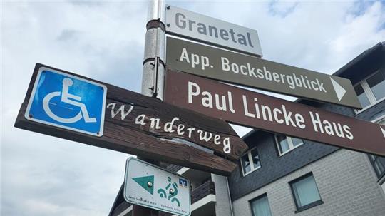 Wegweiser mit den Aufschriften 'Granetal', 'App. Bocksbergblick', 'Paul Lincke Haus' und einem Rollstuhlsymbol mit dem Wort 'Wanderweg' vor einem mehrstöckigen Gebäude und bewölktem Himmel
