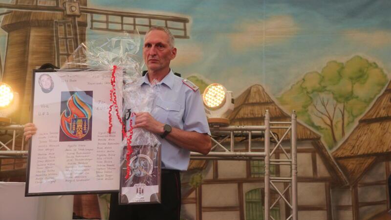Der stellvertretende Ortsbrandmeister Thomas Rademacher hält die Präsente der Ehepartnerinnen und -partner der Feuerwehrleute hoch: Ein Flammenbild mit allen Unterschriften und eine Uhr für die Feuerwache.