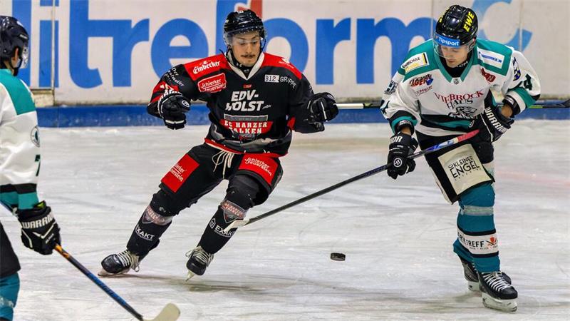 Drei Eishockeyspieler in Aktion auf dem Eis, zwei in weißen Trikots und einer in schwarzem Trikot mit rotem Muster, alle mit Helm und Schläger, Puck auf dem Eis sichtbar