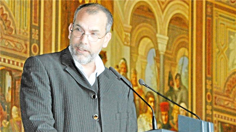 Ein Mann in gestreiftem Anzug steht an einem Rednerpult vor einer Wand mit einem großen, historischen Wandgemälde.