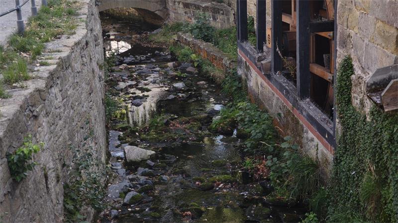 Ein schmaler Bachlauf fließt zwischen alten Natursteinmauern und einem historischen Fachwerkgebäude. Das flache Wasser ist von Steinen, Moos und Laub durchsetzt; im Hintergrund ist eine kleine steinerne Brücke zu sehen.