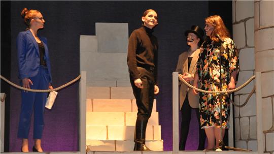 Vier Personen auf einer Bühne mit Kulisse aus gestapelten Kartons, zwei Frauen und zwei Männer, eine Frau trägt einen blauen Anzug, eine andere ein geblümtes Kleid, ein Mann trägt einen schwarzen Rollkragenpullover, der andere einen Hut und einen grauen Blazer