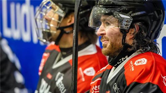 Zwei Eishockeyspieler in schwarzen Helmen und rot-schwarzen Trikots nebeneinander auf der Bank sitzend