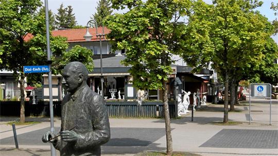 Bronzestatue eines Mannes mit Anzug und Krawatte auf einem Platz mit Bäumen und Gebäuden im Hintergrund