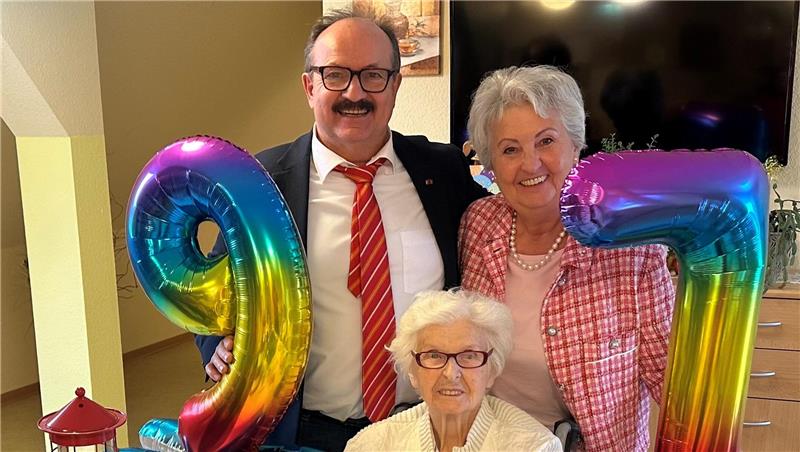 Drei Personen, eine ältere Frau sitzt an einem Tisch mit Blumen, zwei Personen stehen dahinter und halten bunte Luftballons in Form der Zahlen 9 und 7