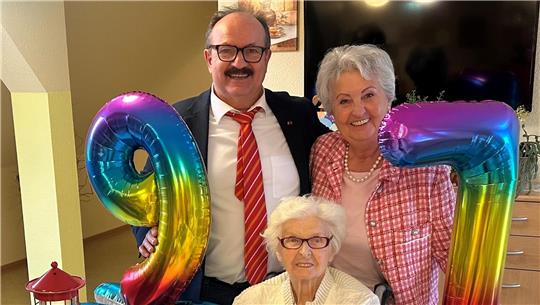 Drei Personen, eine ältere Frau sitzt an einem Tisch mit Blumen, zwei Personen stehen dahinter und halten bunte Luftballons in Form der Zahlen 9 und 7