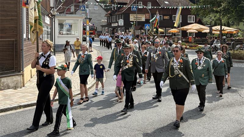 Gruppe von Menschen in Uniformen und mit Abzeichen geht in einem Umzug durch eine Straße in einem Dorf mit Fachwerkhäusern