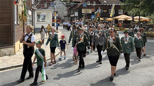 Gruppe von Menschen in Uniformen und mit Abzeichen geht in einem Umzug durch eine Straße in einem Dorf mit Fachwerkhäusern