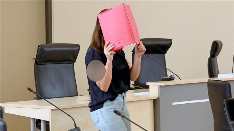 Die Angeklagte betritt den Gerichtssaal mit rotem Aktendeckel vor dem Gesicht.