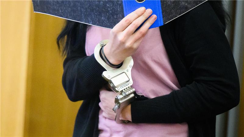 Eine Frau mit schwarzen langen Haaren und einem Aktenordner vor dem Gesicht. Sie trägt Handfesseln an beiden Handgelenken.