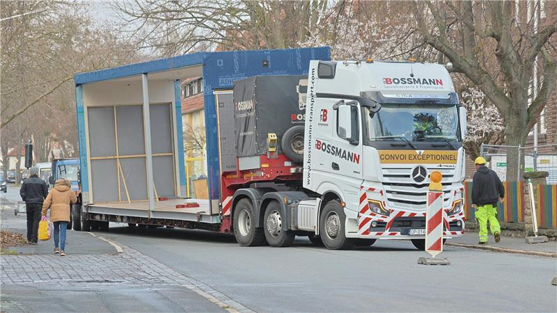 Kran im Einsatz: Spektakulärer Ausbau der Grundschule Oker Sattelzug mit leerem, offenem Auflieger auf einer Straße, daneben zwei Fußgänger und ein Bauarbeiter mit Warnkleidung