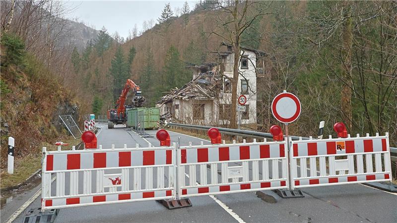 Straßensperre mit rot-weißen Absperrbaken und Warnleuchten vor einem teilweise eingestürzten Haus in bewaldeter Berglandschaft