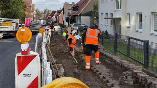 Männer in orange-schwarzen Anzügen arbeiten an einer offenen Straße in einem Wohnviertel.