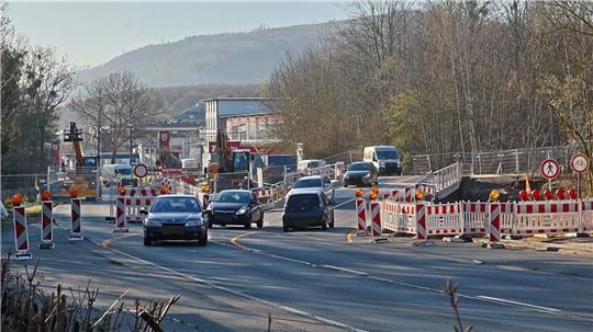 Straße mit mehreren Autos und Baustellenabsperrungen auf beiden Seiten, im Hintergrund Hügel und Bäume
