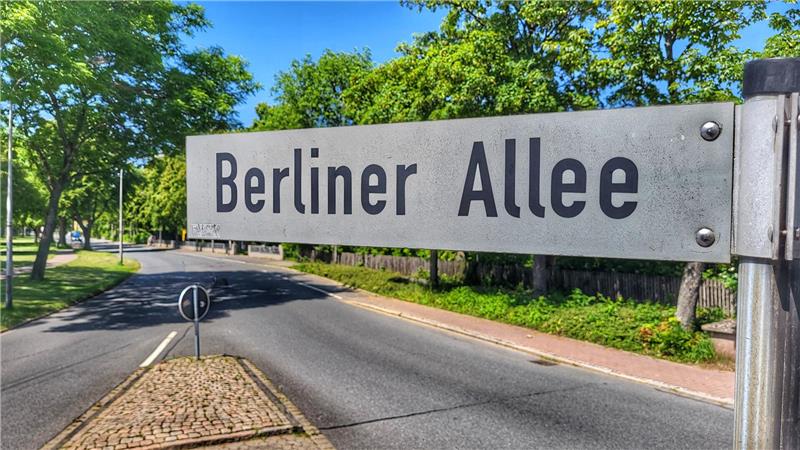 Straßenschild mit der Aufschrift 'Berliner Allee' an einer Straße mit Bäumen und Gehweg