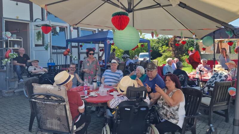 Gruppe älterer Menschen sitzt an mehreren Tischen im Freien unter Sonnenschirmen mit hängender Dekoration in Form von Erdbeeren