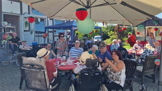 Gruppe älterer Menschen sitzt an mehreren Tischen im Freien unter Sonnenschirmen mit hängender Dekoration in Form von Erdbeeren