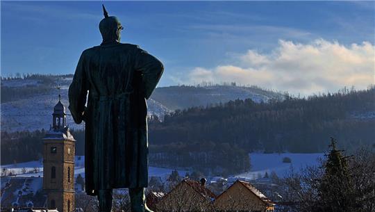 Rückansicht einer Statue mit Pickelhaube vor verschneiter Hügellandschaft und Gebäuden unter blauem Himmel.