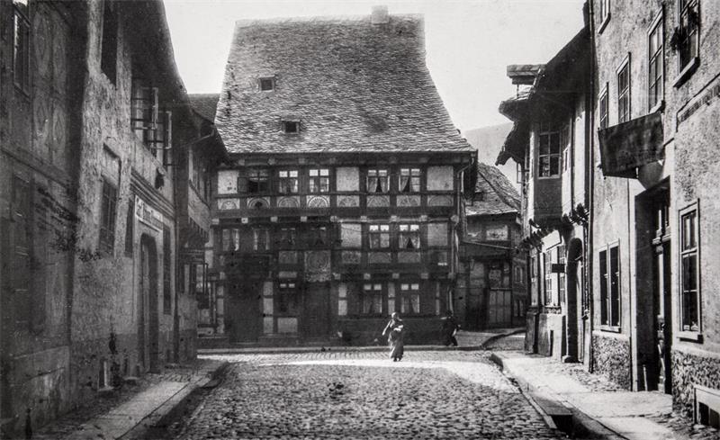 Eine Kopfsteinpflasterstraße, umsäumt von alten Fachwerkhäusern; im Hintergrund gehen einige Menschen die Straße entlang. 
