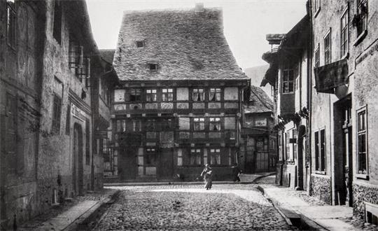 Eine Kopfsteinpflasterstraße, umsäumt von alten Fachwerkhäusern; im Hintergrund gehen einige Menschen die Straße entlang. 