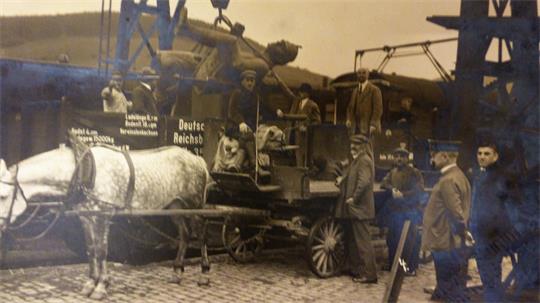 Historisches Schwarz-weiß-Foto mit Pferdefuhrwerk, mehreren Männern in Anzügen und einem Lastwagen mit der Aufschrift 'Deutsche Reichs'