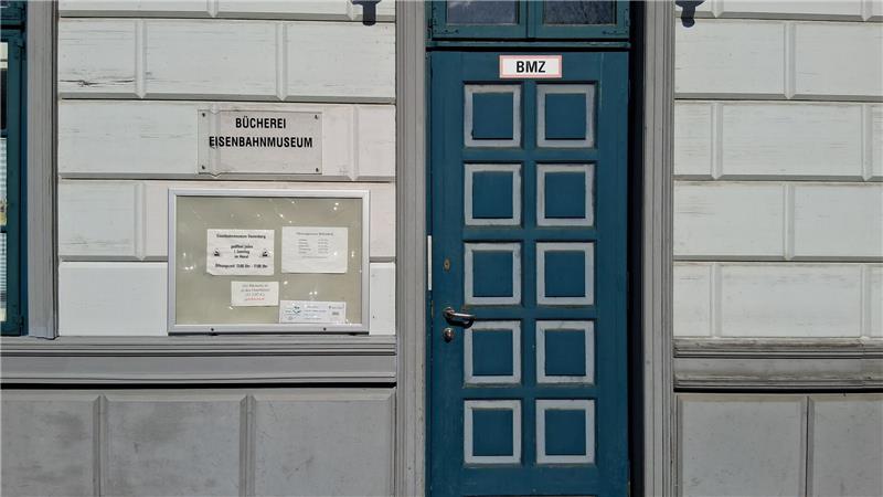 Fassade eines Gebäudes mit einer blauen Tür mit quadratischen Feldern und einem Schild mit der Aufschrift 'BMZ' sowie einem Schaukasten mit mehreren Aushängen.