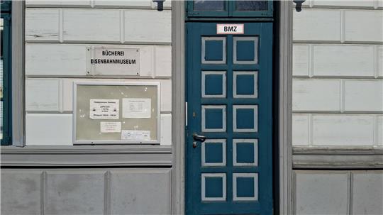 Fassade eines Gebäudes mit einer blauen Tür mit quadratischen Feldern und einem Schild mit der Aufschrift 'BMZ' sowie einem Schaukasten mit mehreren Aushängen.