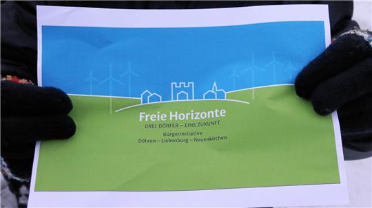Person mit schwarzen Handschuhen hält ein Blatt mit einer stilisierten Landschaft aus grünen Hügeln, blauen Himmel, Windrädern und Gebäuden sowie dem Text 'Freie Horizonte DREI DÖRFER - EINE ZUKUNFT Bürgerinitiative Dürpen - Liesenburg - Neuenkirchen'