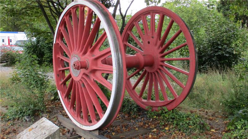 Zwei große rote Eisenräder mit Speichen stehen nebeneinander auf einem Holzpodest im Freien.