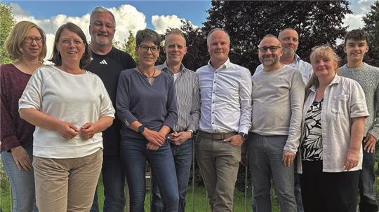 Gruppe von zehn Personen steht im Freien vor grünem Gras und blauem Himmel mit Wolken