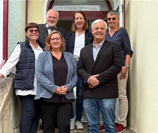 Sechs Männer und Frauen stehen auf einer Treppe mit dem Rücken zur Haustür.