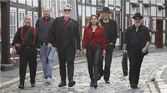 Fünf Personen in schwarzer und roter Kleidung gehen auf einer gepflasterten Straße vor Fachwerkhäusern