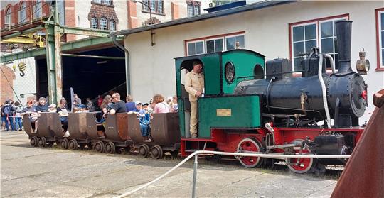 Eisenbahn mit grün-schwarzer Lokomotive und mehreren offenen Waggons, in denen Menschen sitzen, vor einem Industriegebäude mit Backsteinfassade.