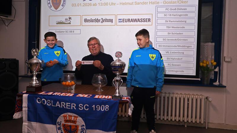 Drei Personen sind vor einer Leinwand mit der Überschrift '38. Fußball-Hallenturnier' und einer Liste von teilnehmenden Mannschaften.