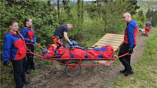Vier Personen in roter und blauer Einsatzkleidung tragen eine Person in einer roten Trage mit einem Rad auf einem Waldweg.