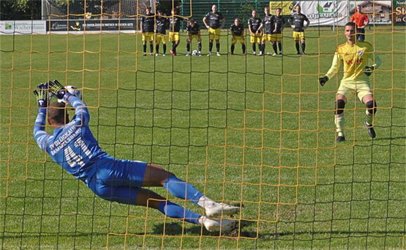 Torwart in blauer Kleidung taucht nach links während ein Spieler in gelber Kleidung einen Elfmeter ausführt und Spieler in schwarzen und weißen Trikots hinter der Mauer stehen