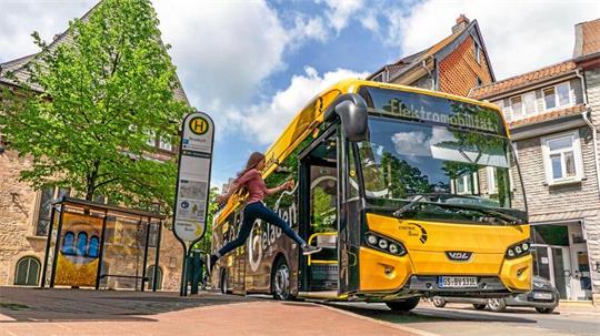 Gelber Elektrobus an Haltestelle, eine Person steigt ein, umgeben von Bäumen und Gebäuden