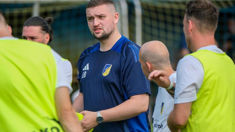 Trainer in blauem Sportshirt mit Vereinslogo spricht mit mehreren Fußballspielern in Trainingsleibchen auf dem Spielfeld