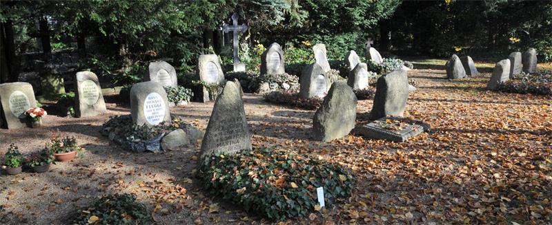 Friedhof mit mehreren alten, unregelmäßig geformten Grabsteinen auf einem mit Laub bedeckten Boden und Bäumen im Hintergrund