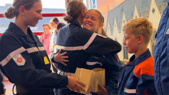 Jugendliche in Feuerwehruniformen umarmen sich und übergeben ein Paket in einem Raum mit gemusterter Wand.