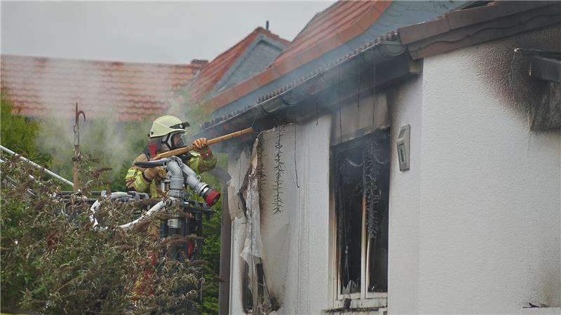 Feuerwehrmann mit Helm und Schutzkleidung löscht an einem brennenden Haus mit beschädigtem Fenster, aus dem Rauch quillt.