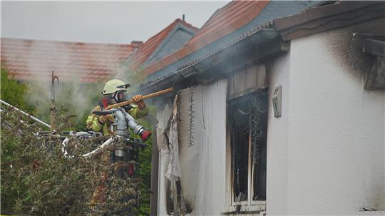 Feuerwehrmann mit Helm und Schutzkleidung löscht an einem brennenden Haus mit beschädigtem Fenster, aus dem Rauch quillt.