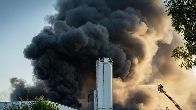 Dichter schwarzer Rauch steigt hinter einem Industriegebäude mit silbernen Silos auf, rechts ist eine Drehleiter im Einsatz.