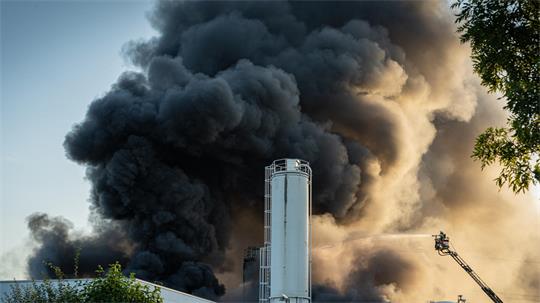 Dichter schwarzer Rauch steigt hinter einem Industriegebäude mit silbernen Silos auf, rechts ist eine Drehleiter im Einsatz.