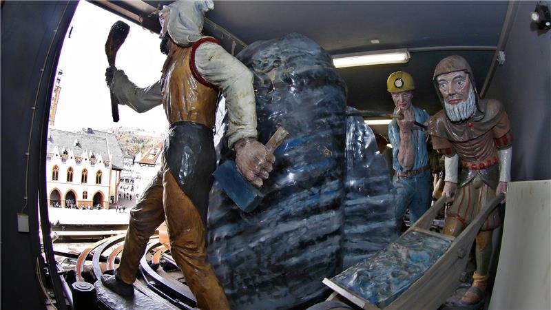 Drei lebensgroße Figuren in historischer Kleidung stehen in einem Raum mit offenem Fenster, eine Figur hält eine Fackel, eine andere steht neben einem Wagen mit blauer Last.