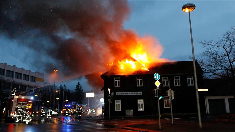 Ein Gebäude steht in Flammen, aus dem Dach schlagen große Feuerflammen und Rauch. Feuerwehrleute und Einsatzfahrzeuge sind vor Ort, es ist Dämmerung.