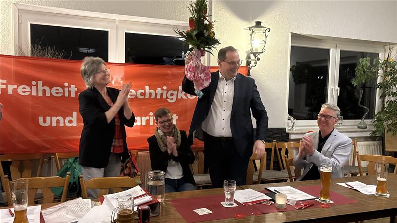 Mann in Anzug hält Blumenstrauß hoch, umgeben von applaudierenden Personen vor rotem Banner mit Text