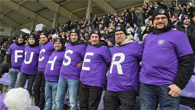 Gruppe von Menschen in lila T-Shirts mit jeweils einem großen weißen Buchstaben auf der Brust, die zusammen das Wort "FUCHSER" bilden, in einem Stadion mit vielen Zuschauern im Hintergrund.