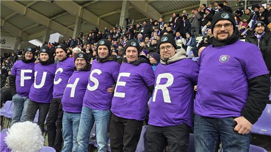 Gruppe von Menschen in lila T-Shirts mit jeweils einem großen weißen Buchstaben auf der Brust, die zusammen das Wort "FUCHSER" bilden, in einem Stadion mit vielen Zuschauern im Hintergrund.
