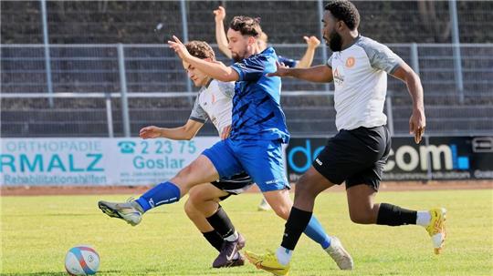 Drei Fußballspieler in Aktion, zwei in weißen Trikots und einer in blauem Trikot, kämpfen um den Ball auf dem Spielfeld.